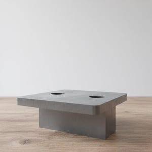 May include: A gray, rectangular concrete candle holder with two circular openings. The candle holder has a square top and a rectangular base. The object is on a wooden surface.