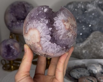May include: A spherical amethyst geode featuring a hollow center filled with jagged, light purple crystal points. The exterior of the stone displays a smooth, rounded surface with natural variations in white and pale pinkish-brown mineral tones.