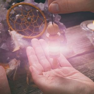 May include: A wooden pendulum with a string hanging over a hand, with a dreamcatcher and amethyst crystals in the background.