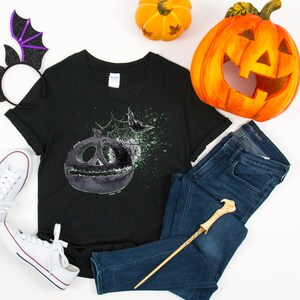 May include: Black t-shirt with a green and gray graphic of a jack-o'-lantern with a spider web and a bat. The shirt is laid out on a white surface with a pair of white sneakers, a purple bat headband, a wooden wand, a pair of blue jeans, and two orange pumpkins.