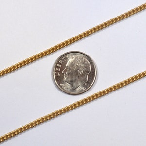 May include: A gold chain necklace with a curb link pattern. The chain is laid out on a white surface next to a US dime for scale.
