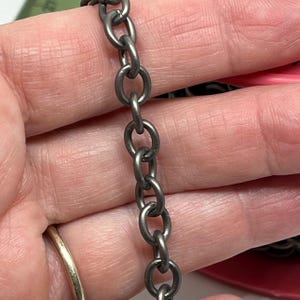 May include: Close-up of a gunmetal-colored chain. The chain is composed of oval links, each connected to the next. The chain is held in a hand, with a gold ring visible in the background. The chain is a jewelry component.