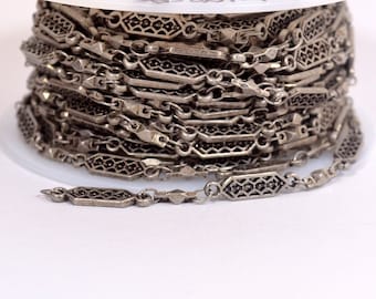 May include: A spool of silver-toned chain with a decorative filigree design. The chain is wound tightly around the spool, and the end of the chain is visible in the foreground.