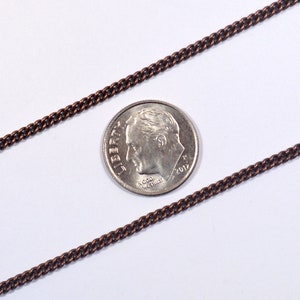 May include: A close-up of a copper chain next to a US dime coin. The chain is about the same width as the coin. The chain is a flat, curb style chain.