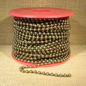 May include: A spool of brass ball chain, wound around a pink plastic spool. The chain is made of small, round brass beads.