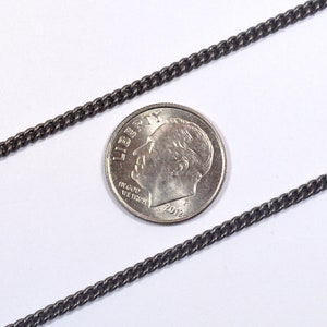 May include: A close-up of a black chain necklace, next to a US dime coin for scale. The chain is made of small, linked metal pieces.
