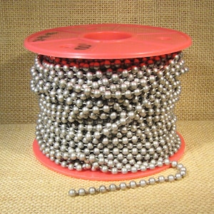May include: A spool of silver ball chain, wound around a red plastic spool. The chain is made of small, round metal beads. The chain is approximately 100 feet long.