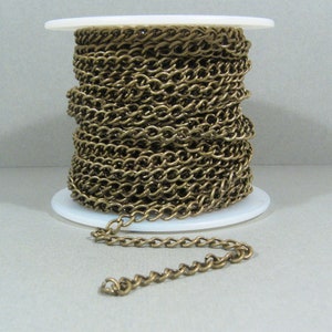 Puede incluir: Un carrete de cadena de latón, enrollado alrededor de un carrete de plástico blanco. La cadena es de estilo curb, con un eslabón plano de forma ovalada. Una pequeña sección de la cadena está desenrollada, mostrando la longitud y el estilo.