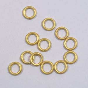 May include: A group of 12 gold jump rings, scattered on a white surface. The rings are all the same size and are made of a shiny metal.