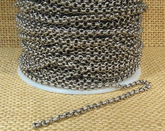 May include: A spool of silver-colored chain with a series of small, circular links. A length of the chain extends from the spool, lying on a tan surface. The chain is likely used for jewelry making or other crafts.