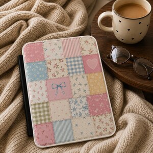 Op de afbeelding: Een rechthoekige e-reader hoes met een patchworkontwerp van diverse vierkanten in zachte pastelkleuren. Het patroon bevat bloemenprints, blauwe strikjes, roze hartjes en ruitjespatronen. De hoes heeft een zwarte rug en een witte rand.