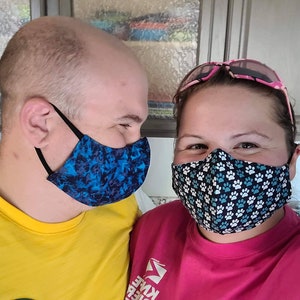 Op de afbeelding: Twee mensen dragen gezichtsmaskers, een met een blauw en zwart gepatroneerd masker en de ander met een zwart masker met witte poot afdrukken. Ze lachen allebei en kijken elkaar aan.