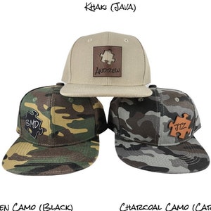 May include: Three baseball caps with different camouflage patterns and leather puzzle pieces with names on them. The caps are khaki, green camo, and charcoal camo.