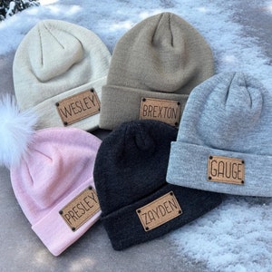 May include: Five knit beanies in different colors with personalized leather patches. The patches have the names "Wesley", "Brexton", "Gauge", "Presley", and "Zayden" on them.