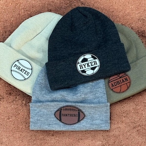 May include: Four knit beanies with different sports-themed patches. The beanies are in various colours, including beige, grey, dark grey, and green. The patches feature a baseball, football, basketball, and rugby ball.