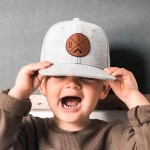 Puede incluir: Gorra de béisbol gris claro con un parche de cuero marrón con las letras "SBL" y un diseño de montaña. La gorra está siendo sostenida por una persona que lleva una camisa marrón de manga larga.