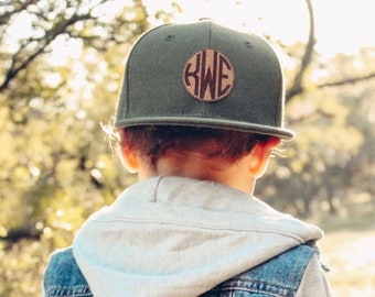 May include: A child wearing a green baseball cap with a brown leather monogram patch that says "KWE". The child is wearing a denim jacket with a grey hoodie underneath.