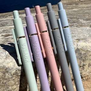 May include: Six pastel-colored pens arranged diagonally. The pens are in shades of green, purple, pink, gray, and blue. Each pen has the word "KRYSTA" printed on the barrel. The pens are resting on a stone surface.