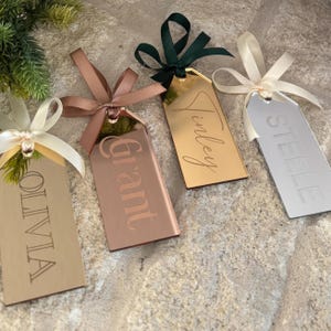 Peut inclure: Quatre étiquettes cadeaux rectangulaires en or, or rose et argent, chacune gravée d'un nom en écriture cursive. Les étiquettes sont ornées de rubans de satin crème, marron et vert foncé. Les noms sont Olivia, Grant, Finley et Steele.