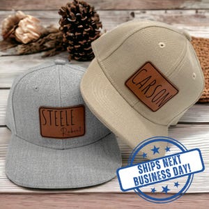May include: A gray baseball cap with a brown leather patch engraved with the name Steele Robert and a tan baseball cap with a brown leather patch engraved with the name Carson. Both caps feature a structured crown and a flat brim design.