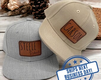 May include: A grey baseball cap with a brown leather patch engraved with the name Steele Robert and a tan baseball cap with a brown leather patch engraved with the name Carson. Both caps feature a structured crown and a flat brim design.
