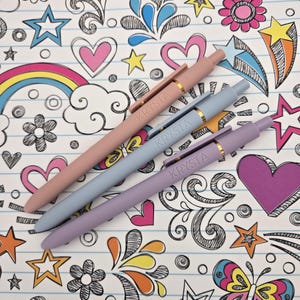 May include: Three pens with the name "Krysta" printed on them. The pens are in shades of pink, blue, and purple. The pens are lying on a sheet of paper with a colorful doodle pattern.