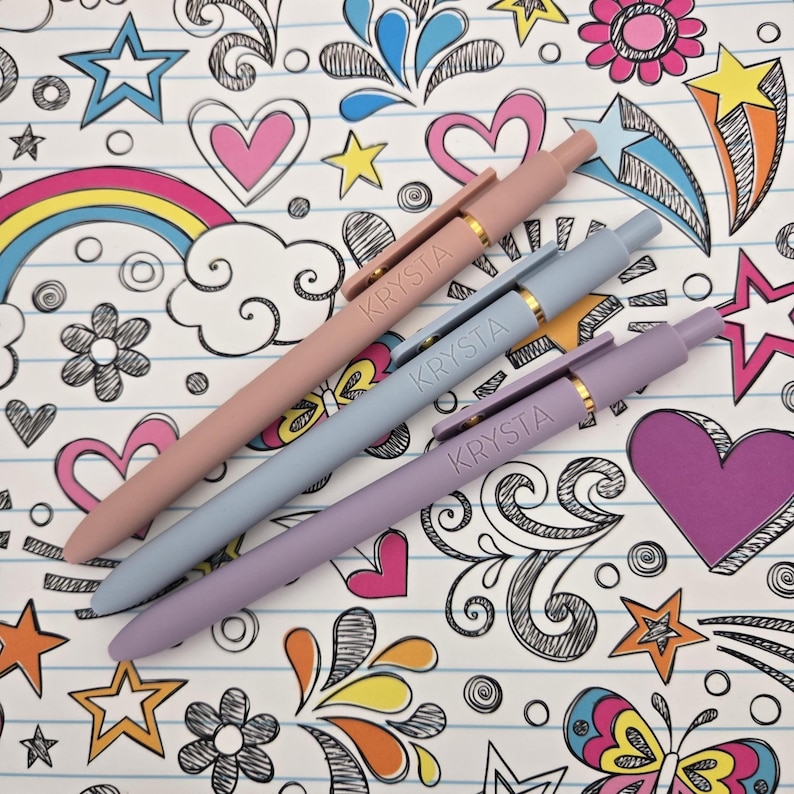May include: Three pens with the name "Krysta" printed on them. The pens are in shades of pink, blue, and purple. The pens are lying on a sheet of paper with a colorful doodle pattern.