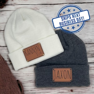 May include: A white knit beanie and a dark gray knit beanie, each featuring a rectangular brown patch. The white beanie patch is engraved with the name KANNYN Ruth, and the dark gray beanie patch is engraved with the name JAXON.