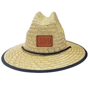 May include: A straw hat with a wide brim and a brown leather patch that reads "COOPER". The hat has a black band around the brim and a black cord around the crown. The hat is made of woven straw.