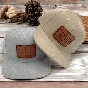 May include: Two baseball caps with personalised leather patches. One cap is light grey with "STEELE Robert" in black lettering. The other is tan with "CARSON" in black lettering. Both caps have a flat peak and a rounded crown.
