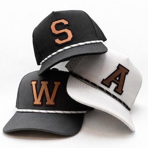 Può includere: Tre cappellini da baseball sovrapposti. Il cappellino superiore è nero con la lettera "S" color rame. Il cappellino centrale è bianco con la lettera "A" marrone. Il cappellino inferiore è grigio scuro con la lettera "W" color rame. Ogni cappellino ha un dettaglio a corda.