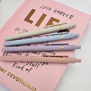 May include: Four pastel-colored pens with gold accents and the word "KRYSTA" printed on each. The pens are arranged on a pink book with the text "LISA HARPER" and "LIF" visible.