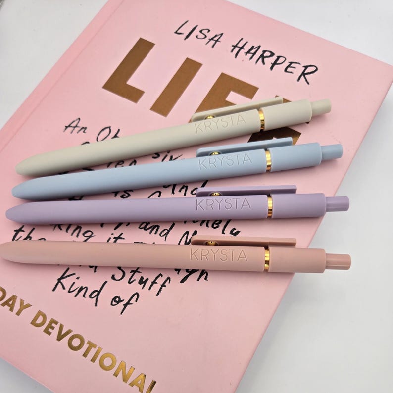 May include: Four pastel-colored pens with gold accents and the word "KRYSTA" printed on each. The pens are arranged on a pink book with the text "LISA HARPER" and "LIF" visible.