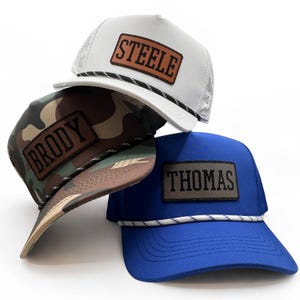 May include: Three baseball caps in white, camouflage, and blue. Each hat features a rectangular leather patch with a name: "STEELE", "BRODY", and "THOMAS". The hats have a curved brim and a rope detail across the front.