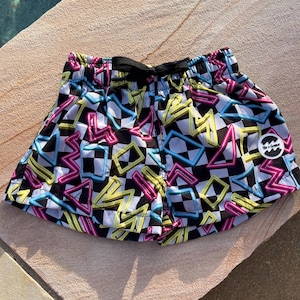 Op de afbeelding: Zwart-wit geblokte zwembroek met neon roze, blauwe en gele geometrische vormen. De broek heeft een zwart trekkoord en een klein logo-patch aan de zijkant.