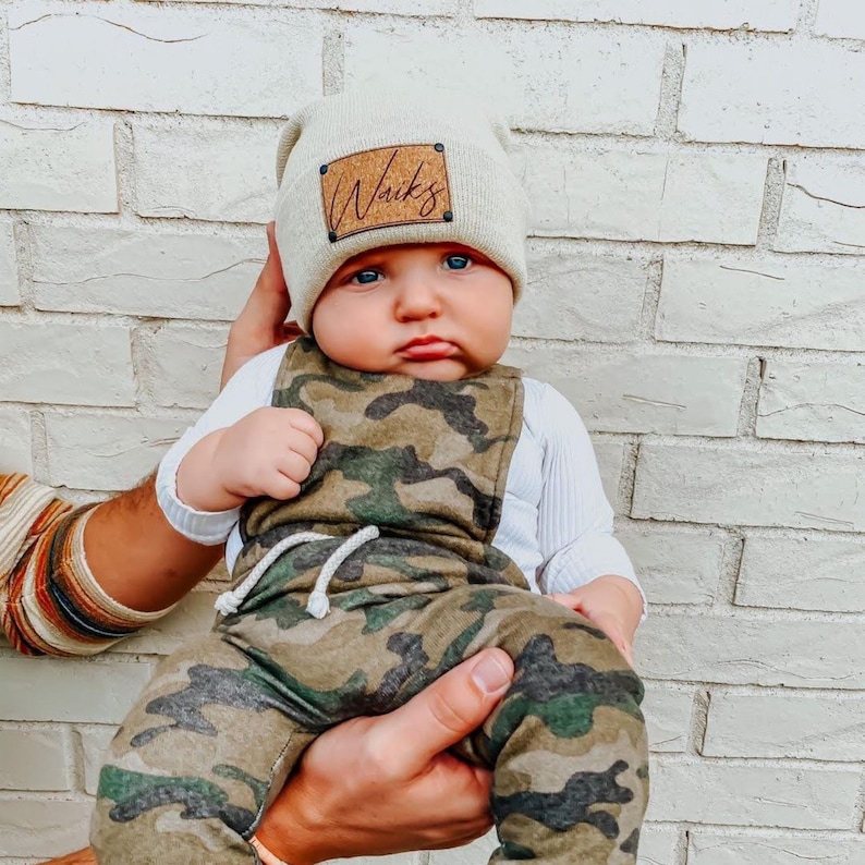 Peut inclure: Un b&eacute;b&eacute; portant un bonnet beige avec le mot "Waits" sur un patch marron, une chemise blanche &agrave; manches longues et une salopette camouflage. Le b&eacute;b&eacute; est tenu, avec un mur de briques blanches en arri&egrave;re-plan.