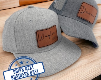 May include: Two grey baseball caps featuring rectangular brown leather patches. The front cap has a flat brim and the text Waylon James. The back cap is a trucker style with a mesh back and a patch with the name Dawson. Both caps are grey.