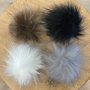 May include: Four faux fur pom poms in different colors: brown, black, white, and gray. The pom poms are arranged in a circle on a wooden surface.