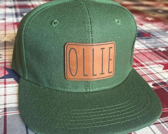 May include: A green baseball cap with a brown leather patch that says "OLLIE" in white lettering.
