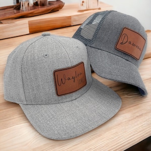May include: Two grey baseball caps with brown leather patches. One cap has the name "Waylon James" and the other has "Dawson" in cursive script. The caps are on a wooden surface.