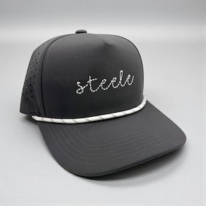 Puede incluir: Una gorra de béisbol gris carbón con la palabra steele bordada en letra cursiva blanca en el panel frontal. La gorra incluye un detalle de cuerda blanca en la base de la corona y paneles laterales perforados para ventilación.