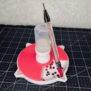 May include: A white and red craft organizer with a clear plastic bottle and a rhinestone-studded pen. The organizer has a tray with compartments holding red and black rhinestones. The pen is black and silver with a pink tip.