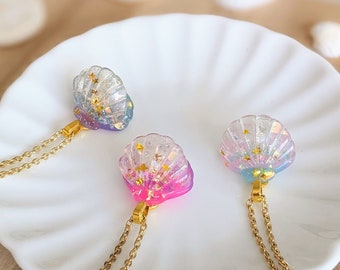 May include: Three gold chain necklaces with iridescent seashell pendants. Each seashell is filled with gold glitter and has a different color gradient. The shells are light blue, pink, and purple.