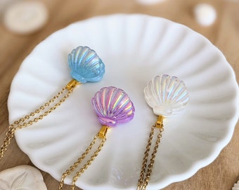 May include: Three seashell pendant necklaces in blue, purple, and white, each with a gold-colored chain. The seashell pendants are iridescent and sit on a white plate. The background includes sand dollars and a wooden surface.