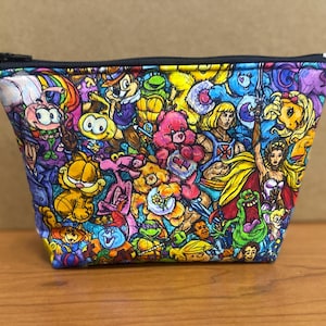 May include: A colorful zippered pouch with a variety of cartoon characters and images printed on it. The characters include Garfield, Care Bears, He-Man, and Teenage Mutant Ninja Turtles.