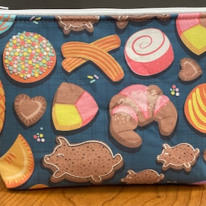 May include: A zippered pouch with a dark blue background and a repeating pattern of various baked goods, including cookies, pastries, and churros. The zipper is silver, and the pouch is made of fabric.