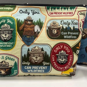 May include: Three zippered pouches with Smokey Bear designs. One is green with a repeating pattern of Smokey Bear's face. The second is cream-colored with Smokey Bear and text that reads "Only You Can Prevent Wildfires." The third is blue with a repeating Smokey Bear pattern.