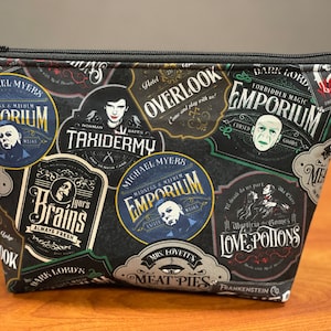 May include: A black zippered pouch with a colorful print of various horror movie themed labels. The labels feature text such as "Overlook Hotel", "Taxidermy", "Brains", "Emporium", "Love Potions", and "Meat Pies".