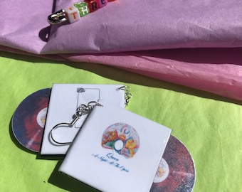 May include: A pair of earrings featuring a Queen album cover, "A Night at the Opera", with a silver chain and a CD-shaped charm. The earrings are on a green surface.