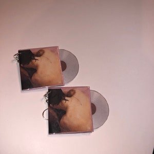 May include: Two silver keychains with a small, round, silver charm. Each keychain features a square, pink-toned card with a nude image of a person. The card is slightly open, revealing a silver disc.